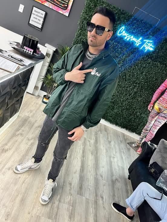 Man in sunglasses and a green jacket pointing at his chest while standing in a tattoo studio.